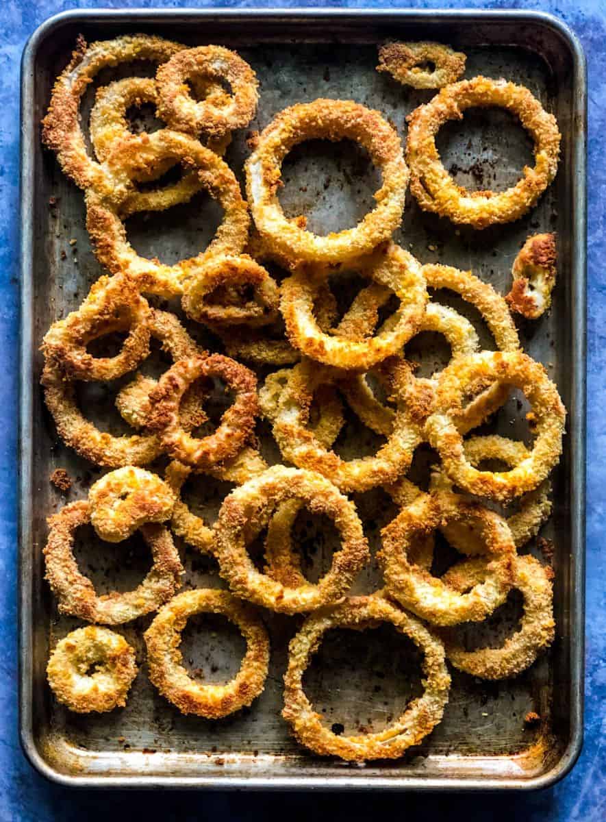 Crispy, golden-brown breaded onion rings are spread across a metal baking sheet placed on a blue textured background.
