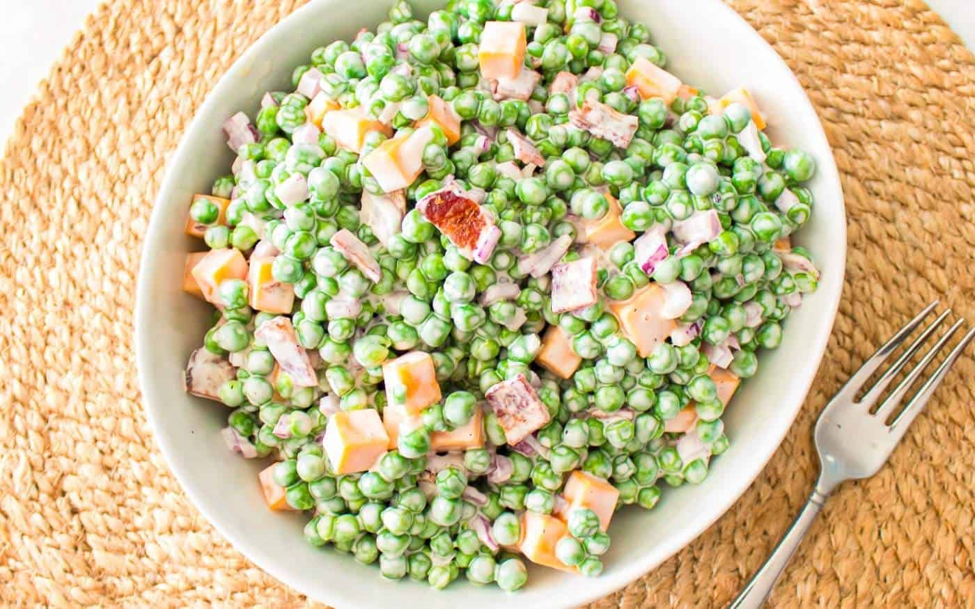 A bowl of creamy pea salad sits on a woven placemat next to a silver fork.