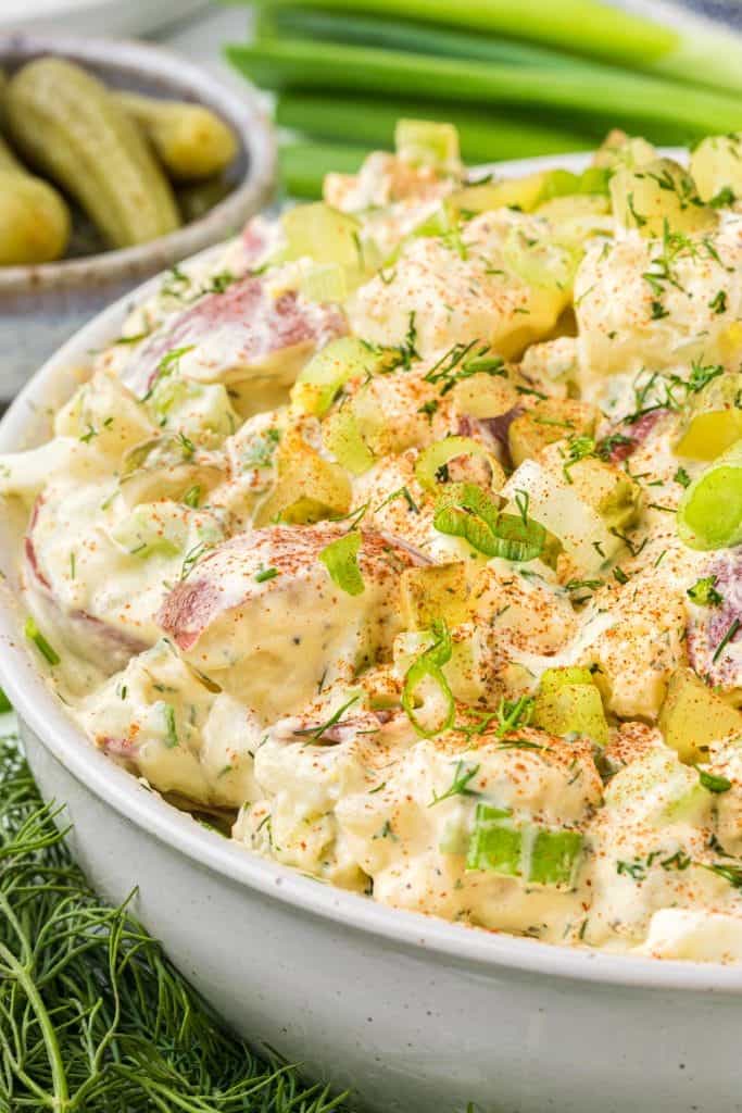 Creamy potato salad with red potatoes, dill pickles, green onions, and fresh dill in a white serving bowl.