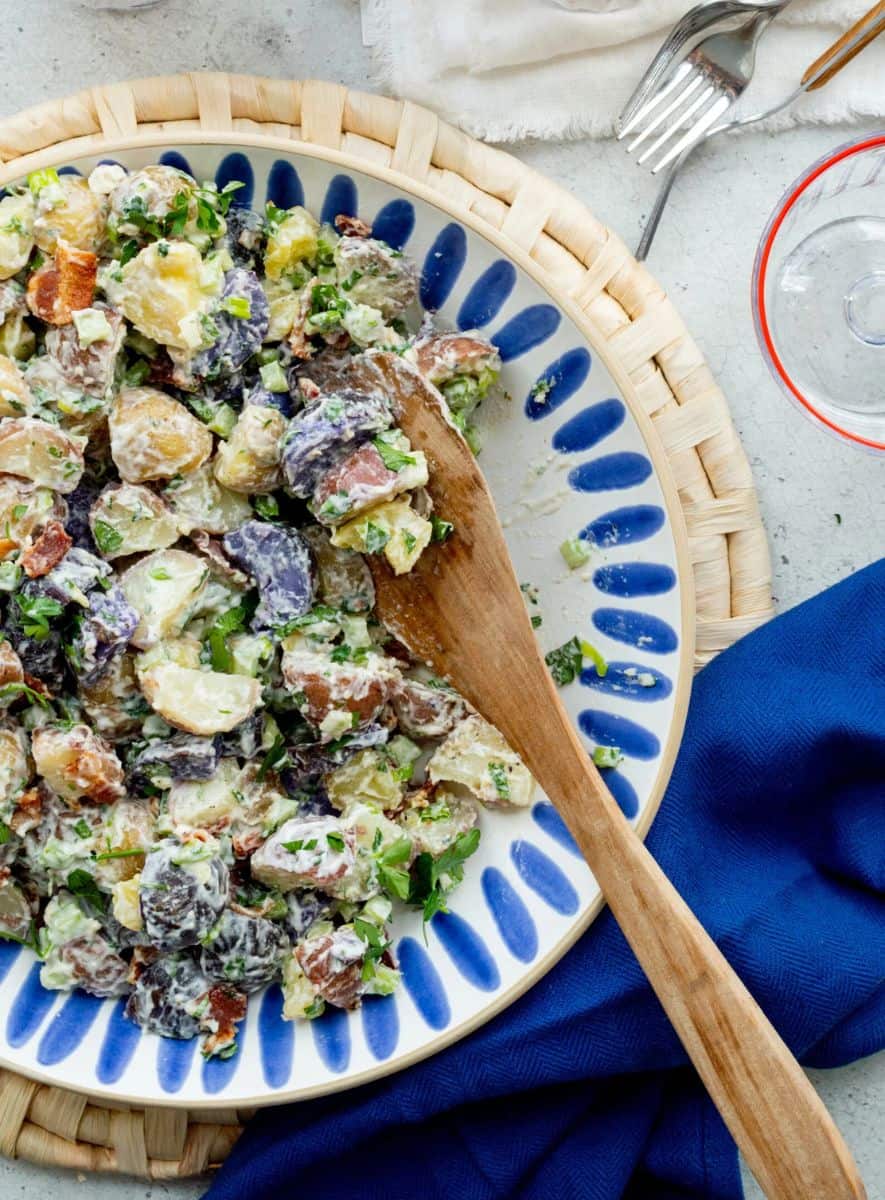 Creamy potato salad with red, white, and purple potatoes, bacon, herbs, and cheese on a serving platter with a wooden spoon.