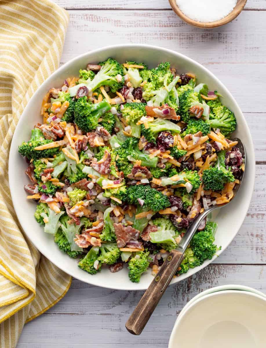 A bowl of broccoli salad with shredded cheese, bacon, red onion, dried cranberries, and pecans, with a spoon and a yellow striped napkin beside it.