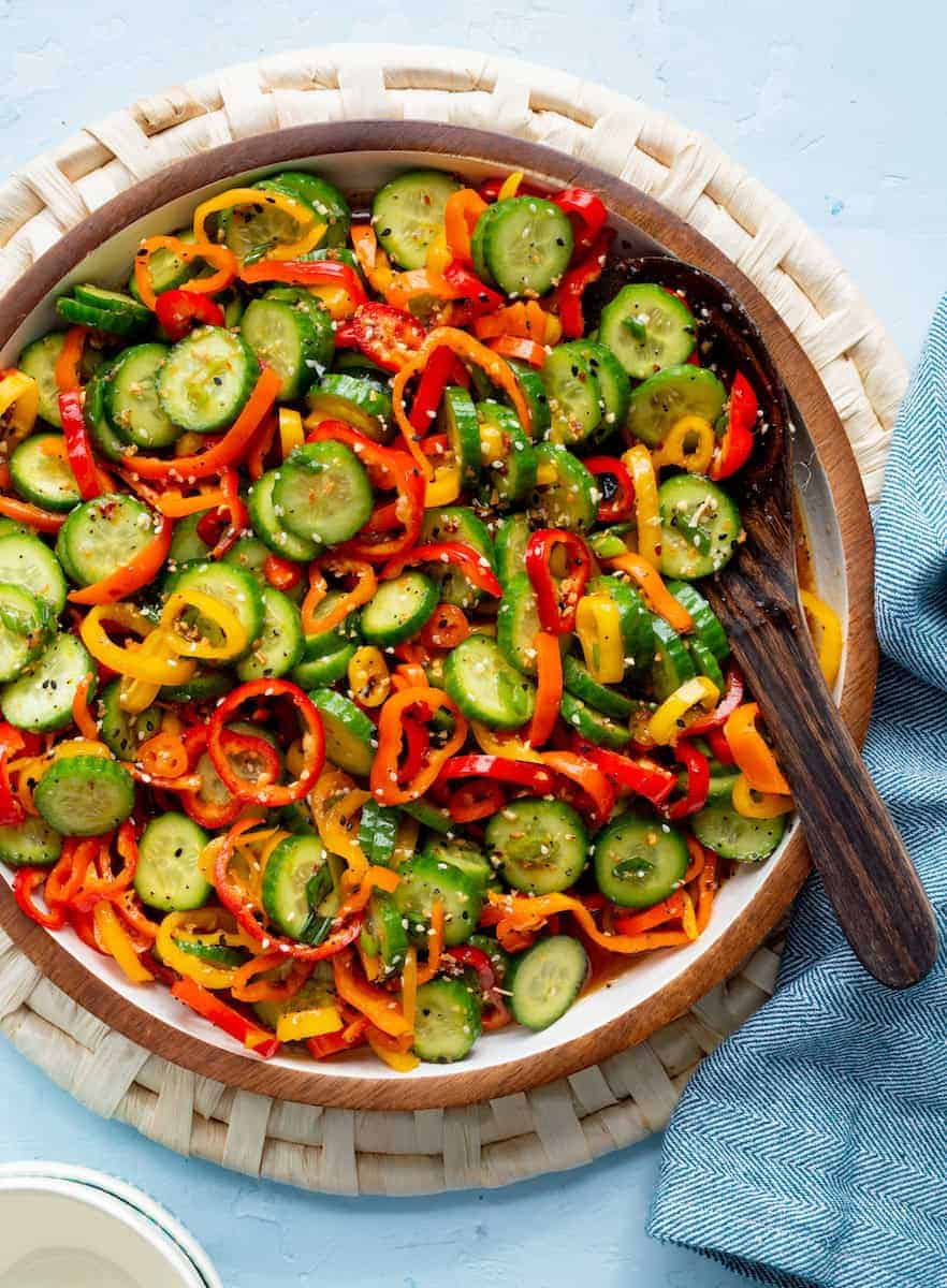 A wooden bowl filled with a colorful salad of sliced cucumbers and red, yellow, and orange bell peppers, with a serving spoon.