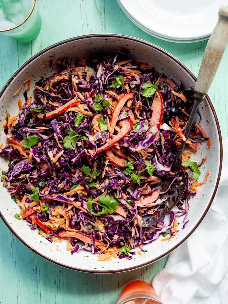 Red cabbage slaw with shredded carrots, sliced red peppers, and fresh cilantro in a large serving bowl with a serving spoon.