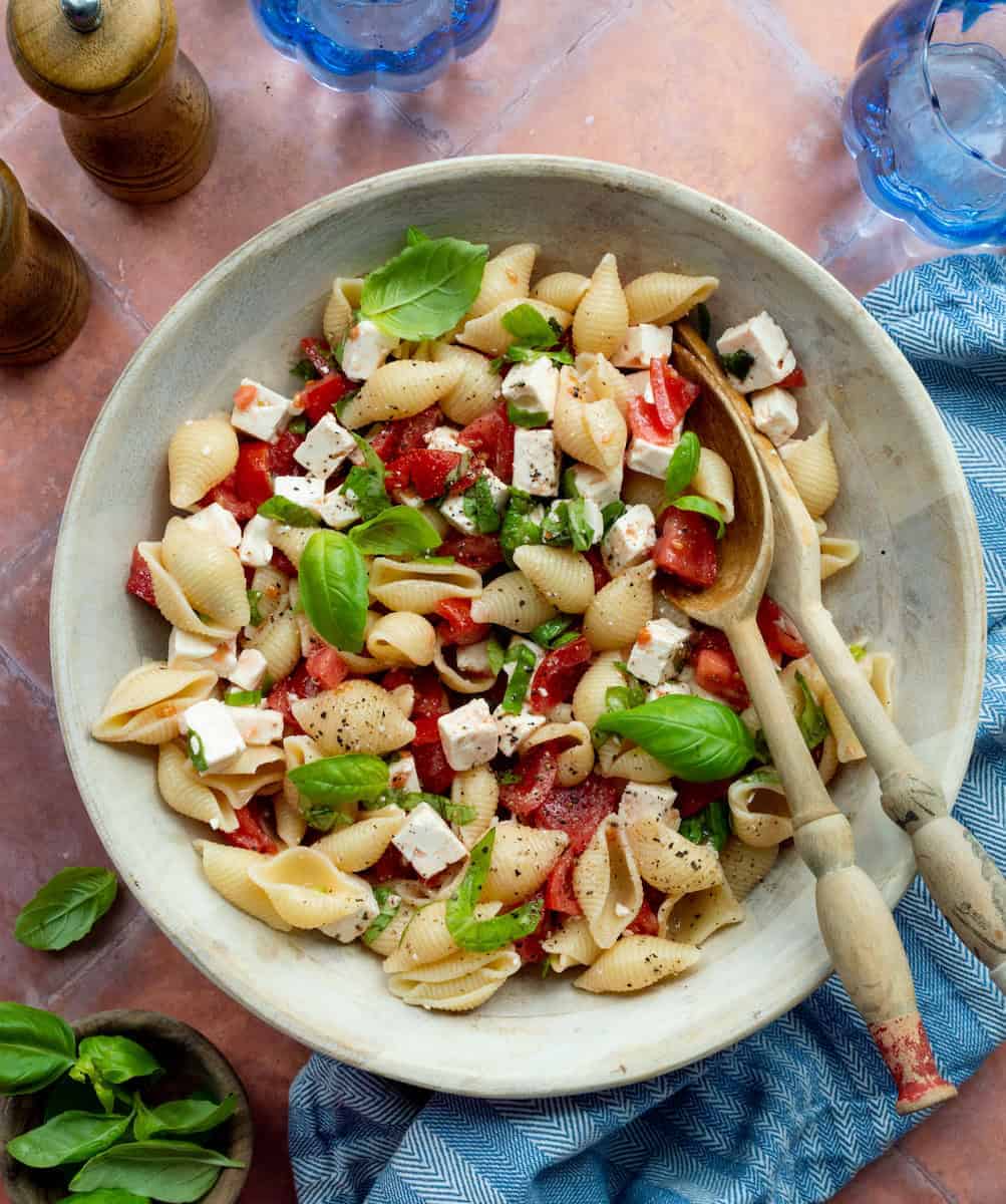 Tomato and feta pasta salad served in a large bowl with fresh basil leaves and wooden serving spoons.