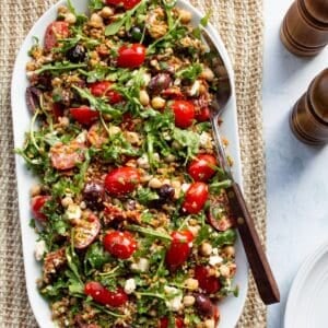 A white oval platter topped with Mediterranean farro salad on a woven linen runner with wooden salt and pepper shakers.
