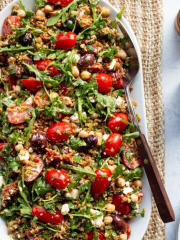 A white oval platter topped with Mediterranean farro salad on a woven linen runner with wooden salt and pepper shakers.