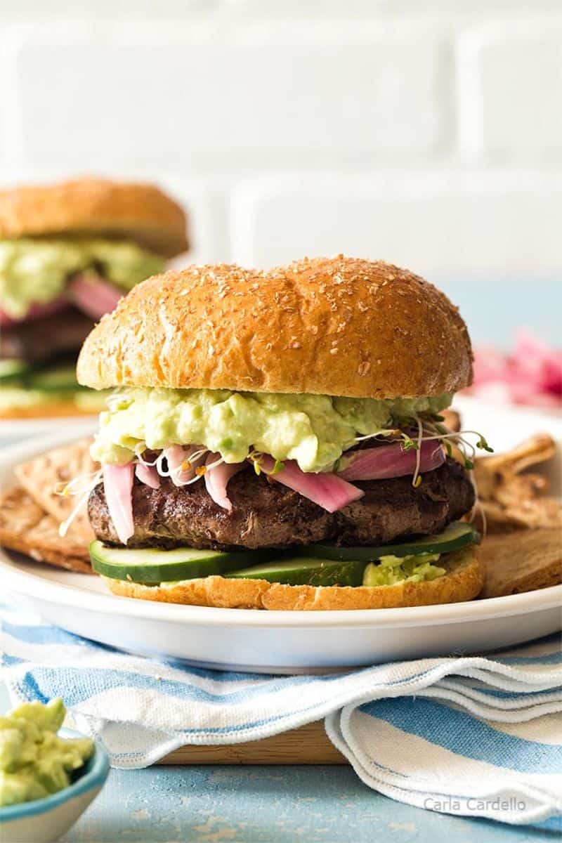Avocado burgers served on buns with burger patties, sliced cucumber, pickled onions, and a chunky avocado topping.