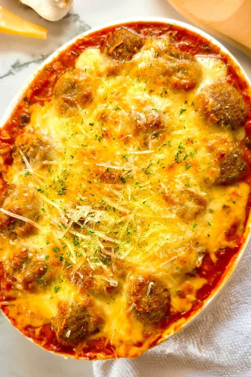 Baked spaghetti and meatballs in a baking dish topped with melted cheese and a sprinkling of herbs.