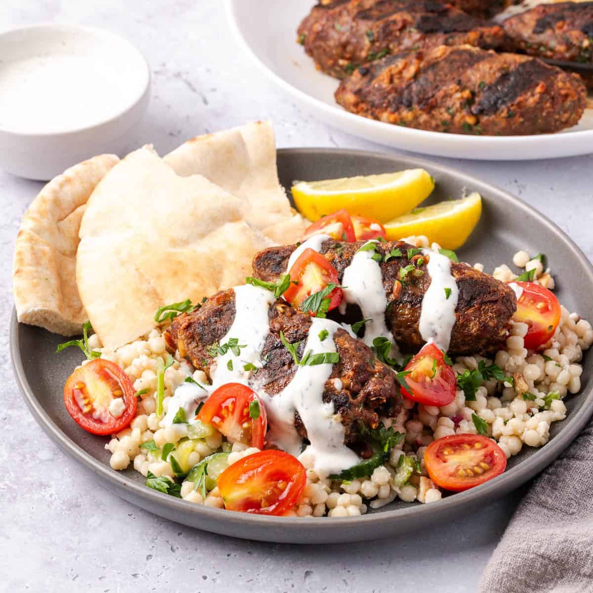 Beef kofta kebabs served with couscous, tomatoes, pita bread, lemon wedges, and a creamy sauce.