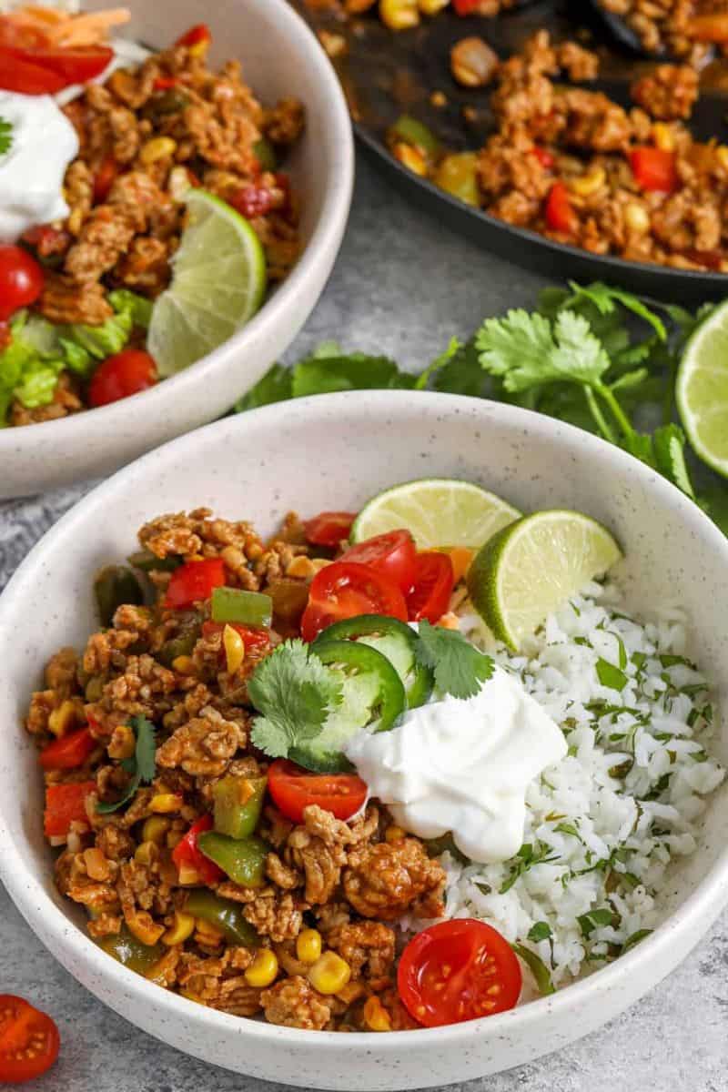 Ground turkey burrito rice bowls with rice, seasoned turkey, corn, peppers, tomatoes, sour cream, jalapeños, lime wedges, and cilantro.