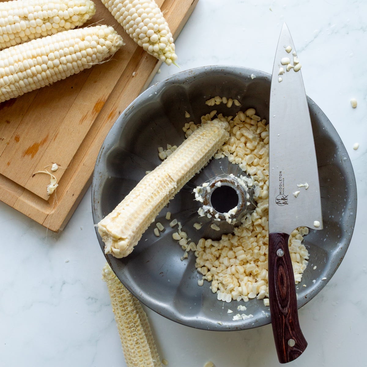 Cutting fresh corn from the cob into a Bundt pan with a cutting board of shucked corn on the side.