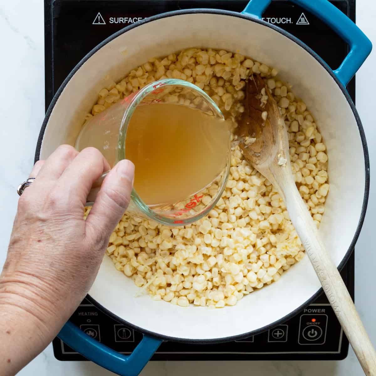 Pouring chicken broth into a blue-handled Dutch oven of fresh corn kernels.