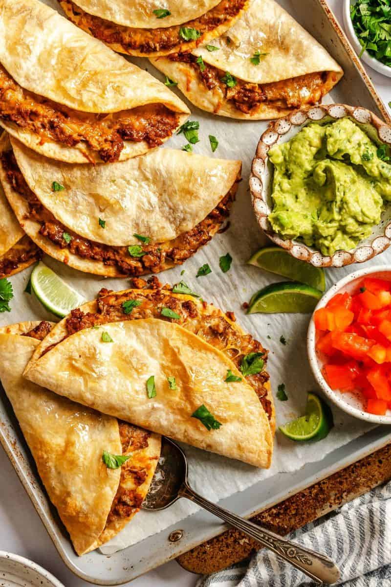 Baked ground beef and bean tacos with crispy folded tortillas, served with guacamole, diced tomatoes, lime wedges, and chopped herbs.