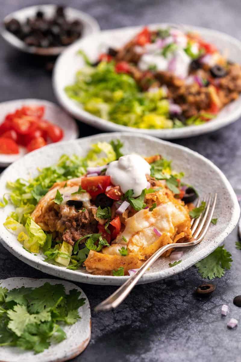 Slow cooker enchilada casserole served with lettuce, tomatoes, black olives, red onion, sour cream, and chopped herbs.