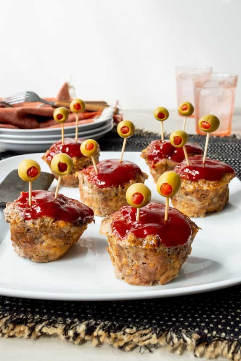 Mini monster meatloaf topped with red sauce and green olives on toothpicks for a playful Halloween presentation.
