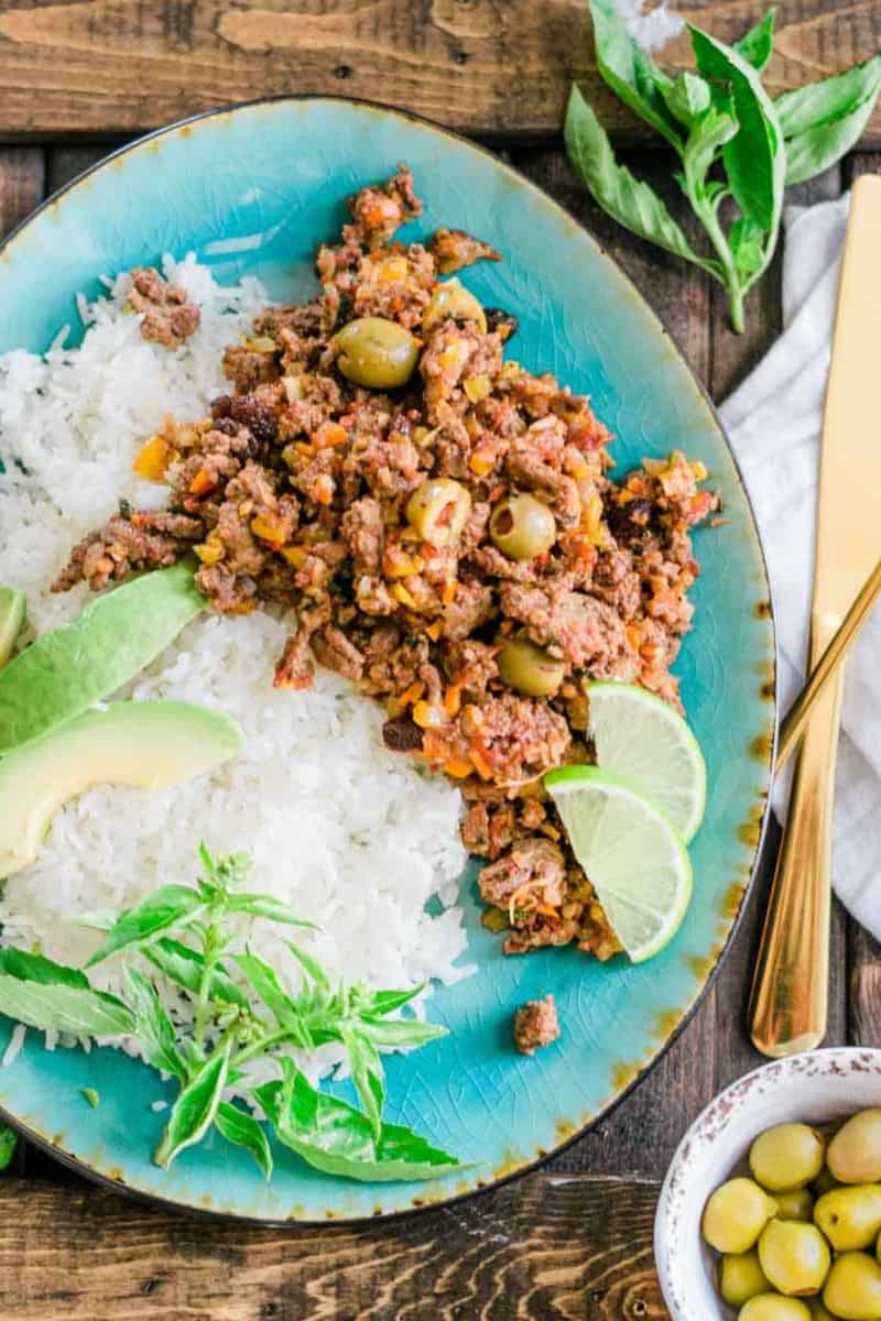 Puerto Rican picadillo served with white rice, avocado slices, green olives, lime wedges, and fresh herbs.