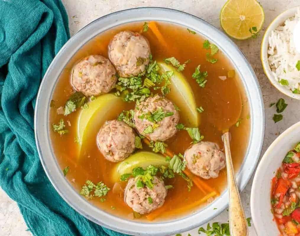 Sopa de albondigas with meatballs and vegetables in a clear broth, topped with chopped herbs.