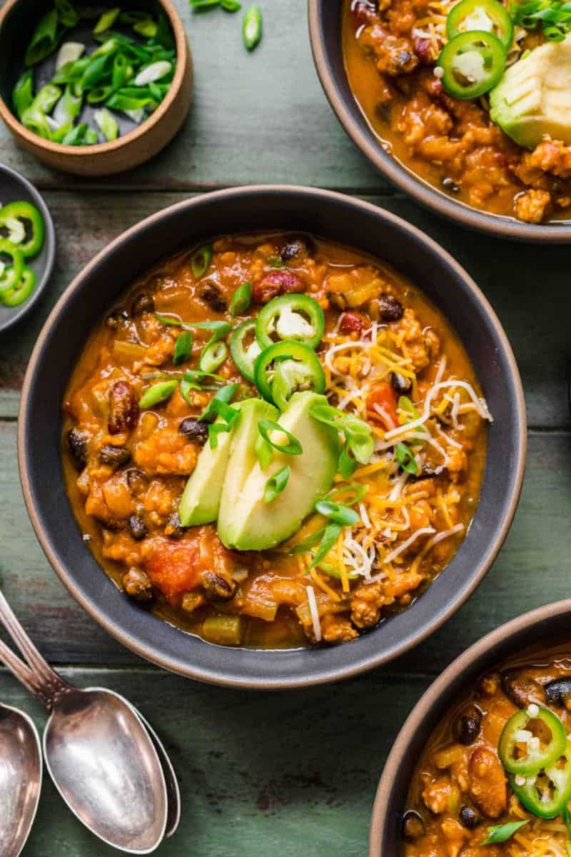 Turkey pumpkin chili served in a bowl with beans, avocado slices, jalapeños, shredded cheese, and green onions.