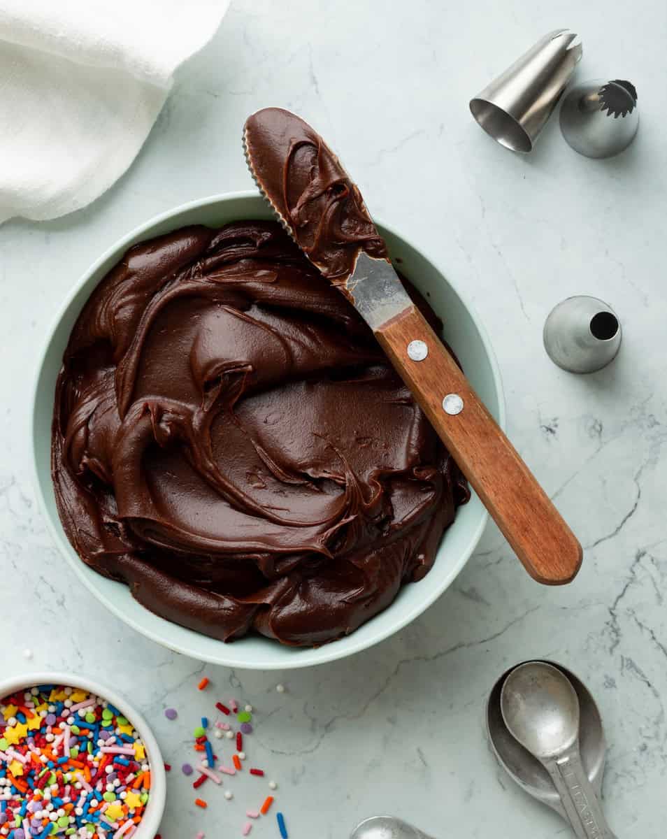 A light green bowl of chocolate fudge frosting surrounded by pastry tips, a bowl of sprinkles, and measuring spoons.