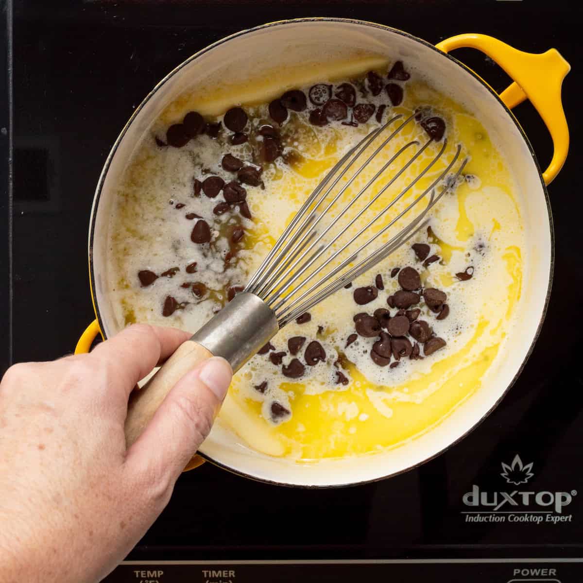 Whisking together melting butter, cream, and chocolate chips in a yellow saucepan.