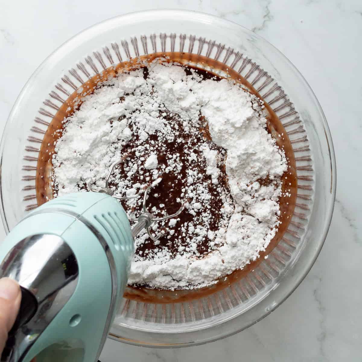 Beating powdered sugar into a chocolate mixture in a glass bowl with an aqua hand mixer.