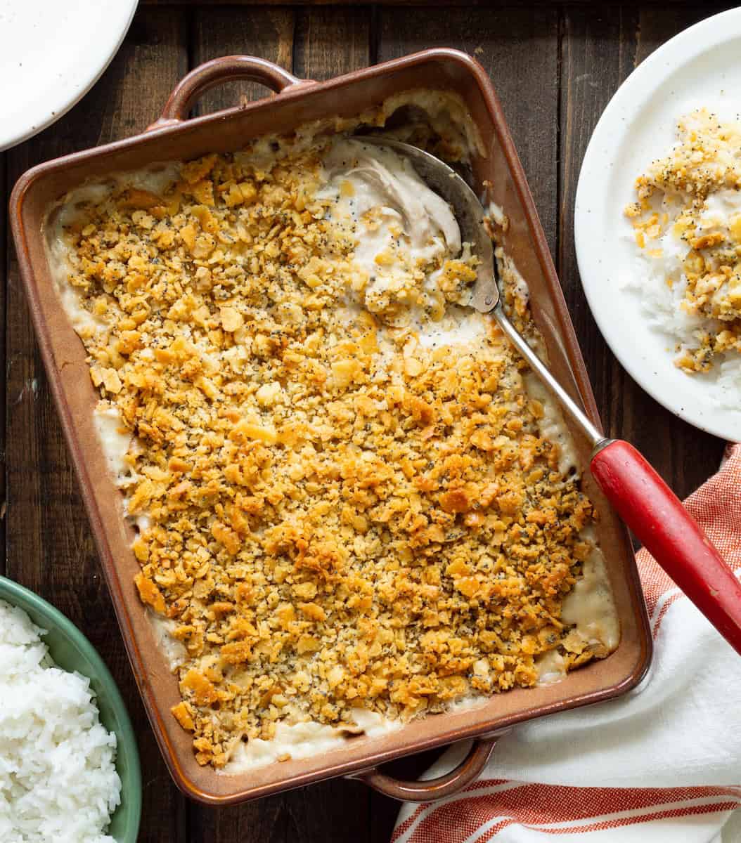 Poppyseed chicken in a brown casserole dish with a red handled spoon and serving to the side.