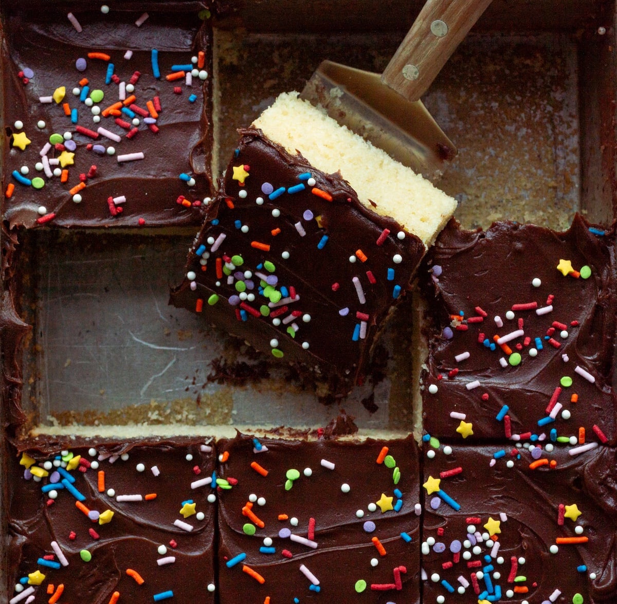 A square of yellow cake with chocolate frosting being lifted out of a metal pan.