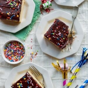 Slices of yellow sheet cake with chocolate frosting topped with sprinkles on small octagonal plates surrounded by party blowers.