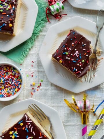 Slices of yellow sheet cake with chocolate frosting topped with sprinkles on small octagonal plates surrounded by party blowers.