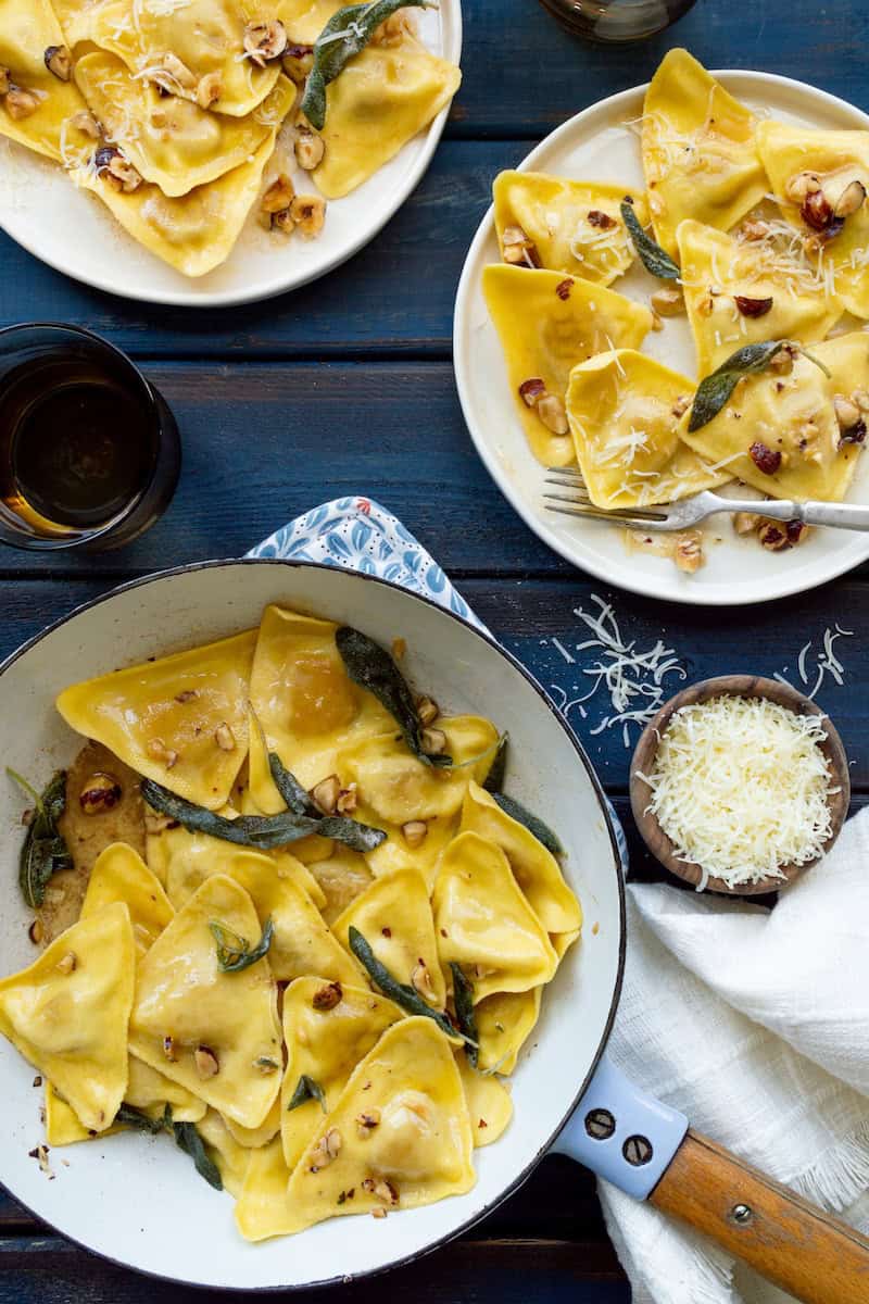 Butternut squash ravioli with sage butter, topped with toasted nuts, fresh sage leaves, and grated parmesan cheese, served on white plates.