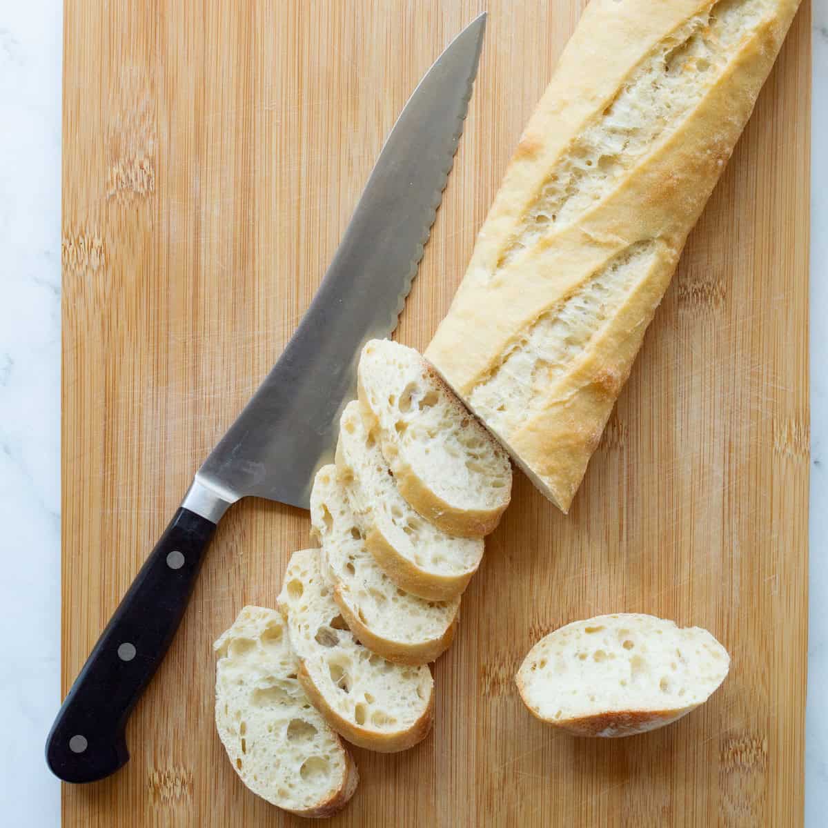 Cutting a baguette into thin slices on a wooden board with a serrated knife.