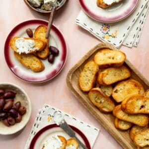 Homemade garlic crostini on a rectangular wooden tray with small plates topped with olives and goat cheese spread on crostini.