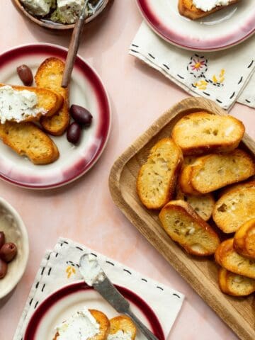 Homemade garlic crostini on a rectangular wooden tray with small plates topped with olives and goat cheese spread on crostini.