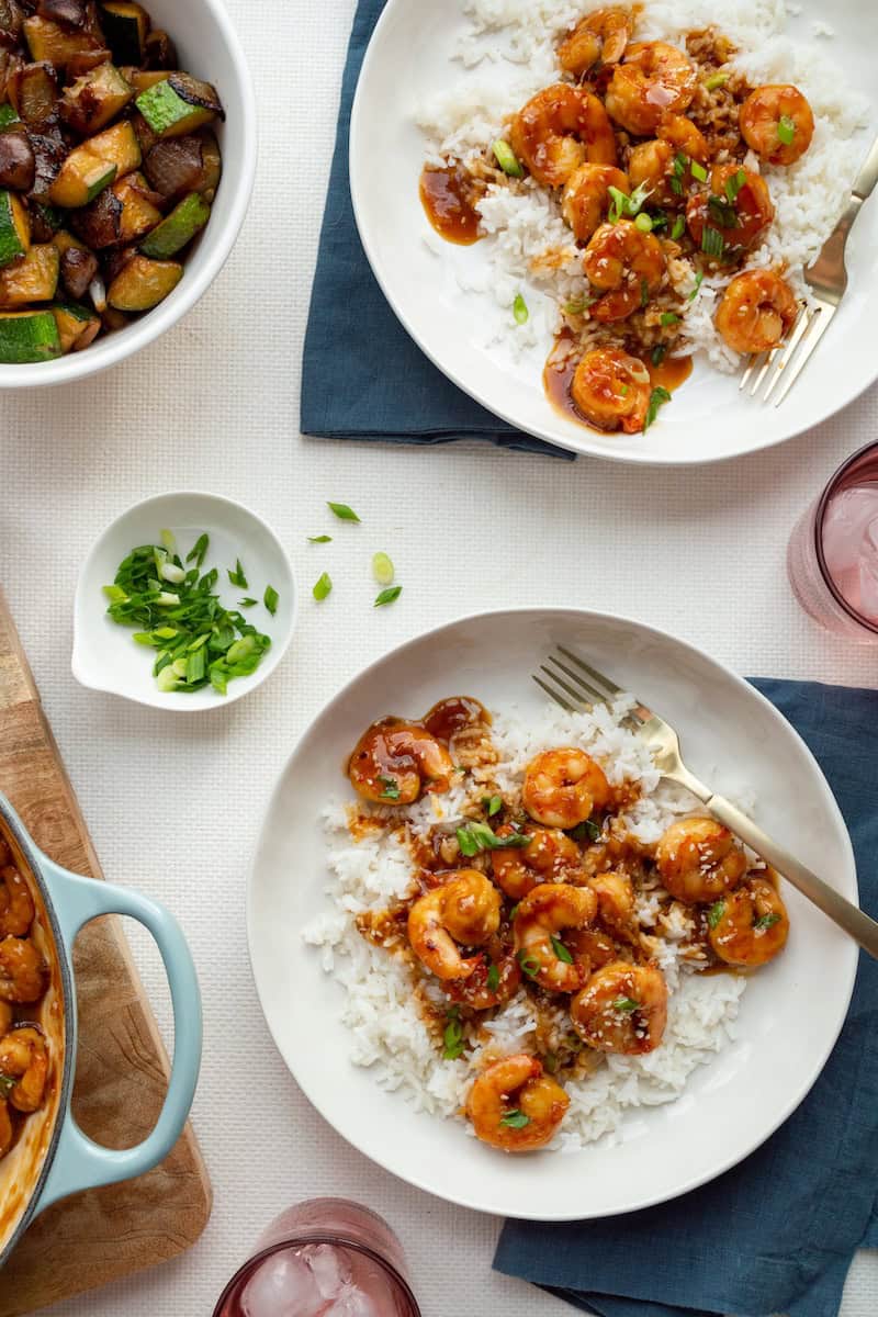 Honey garlic shrimp coated in a sticky sauce, served over white rice with sesame seeds and green onions, alongside sautéed vegetables.
