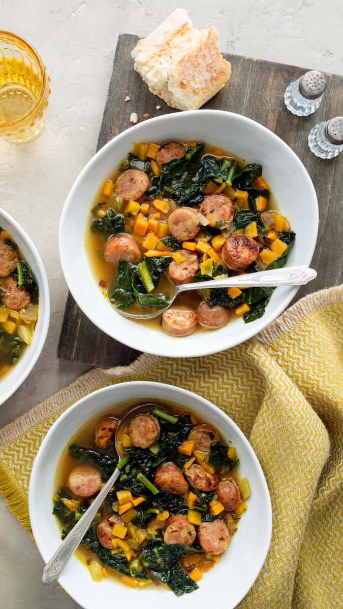 Trader Joe's holiday hash and sausage stew with sliced sausage, diced vegetables, and leafy greens in a savory broth, served in white bowls.
