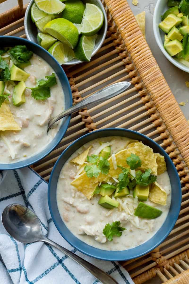 Instant Pot white bean and turkey chili topped with avocado, shredded cheese, tortilla chips, and cilantro, served in blue bowls with lime wedges.