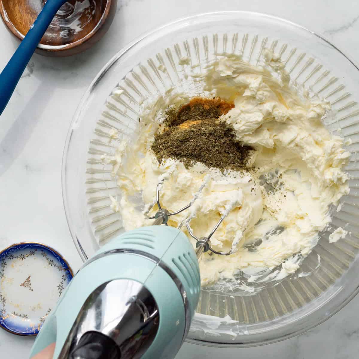 Blending cream cheese, sour cream and spices in a glass bowl with an aqua hand mixer.