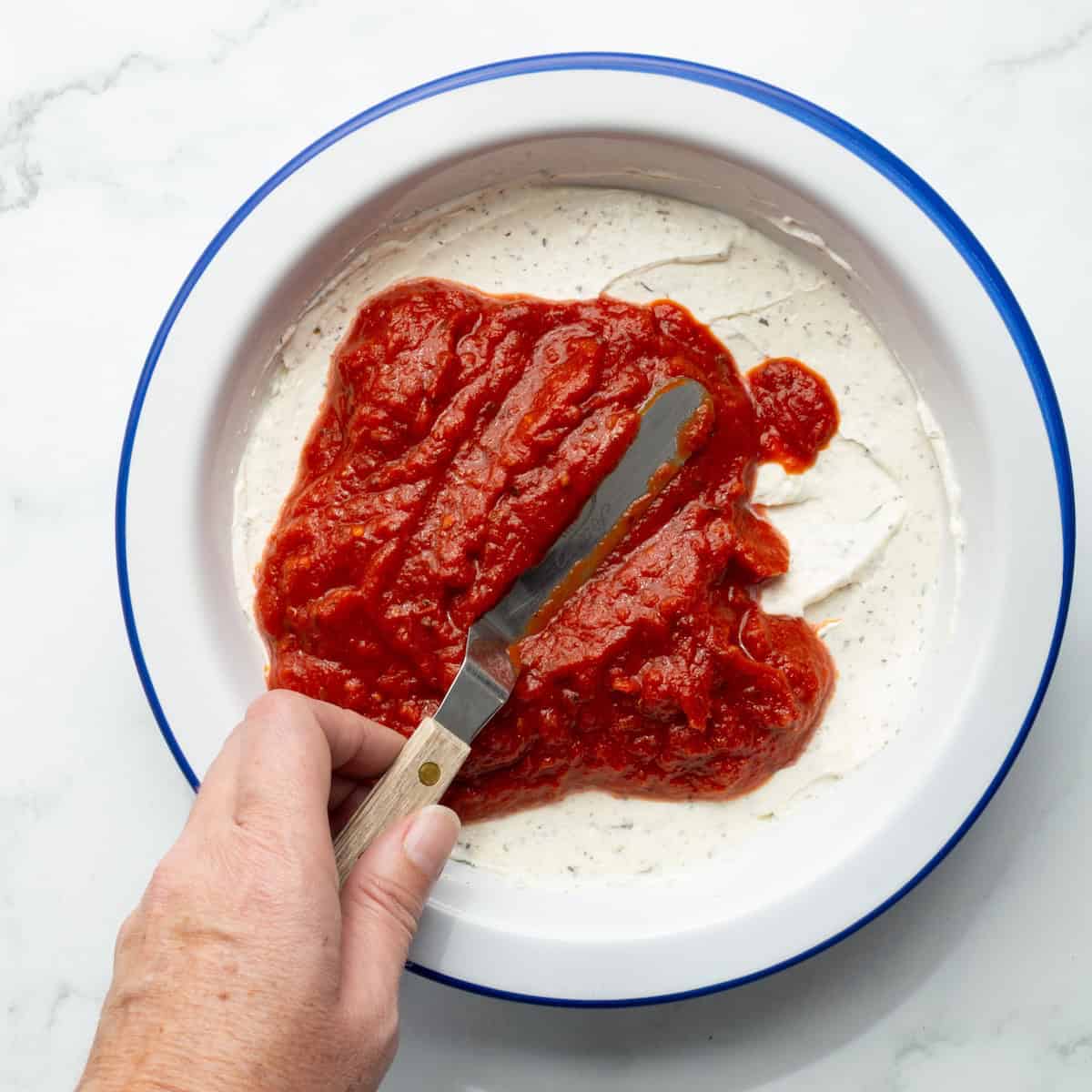 Spreading marinara sauce over the cream cheese mixture in a round baking dish.