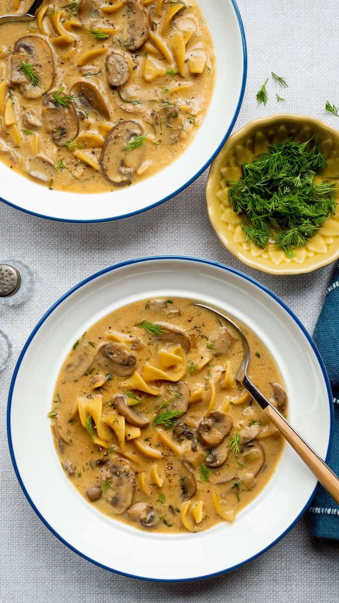 Polish mushroom soup recipe with sliced mushrooms, egg noodles, and fresh dill in a creamy broth, served in white bowls with blue rims.