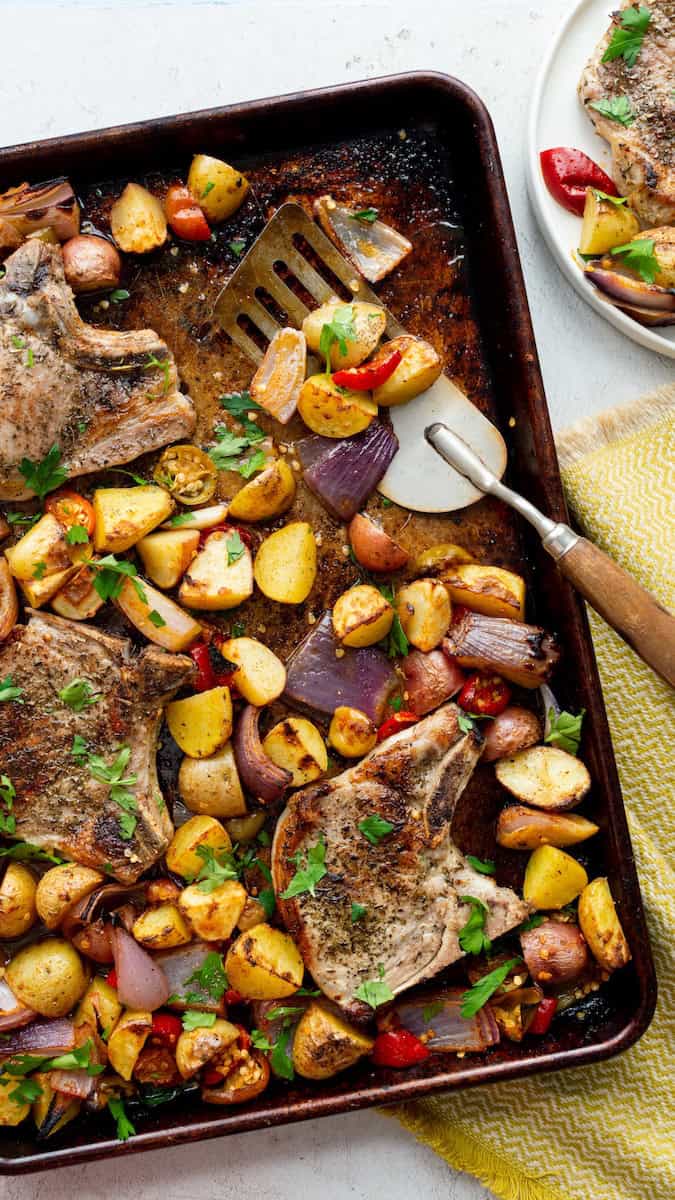 Pork chops with cherry peppers, roasted potatoes, and red onions on a sheet pan, garnished with fresh parsley.