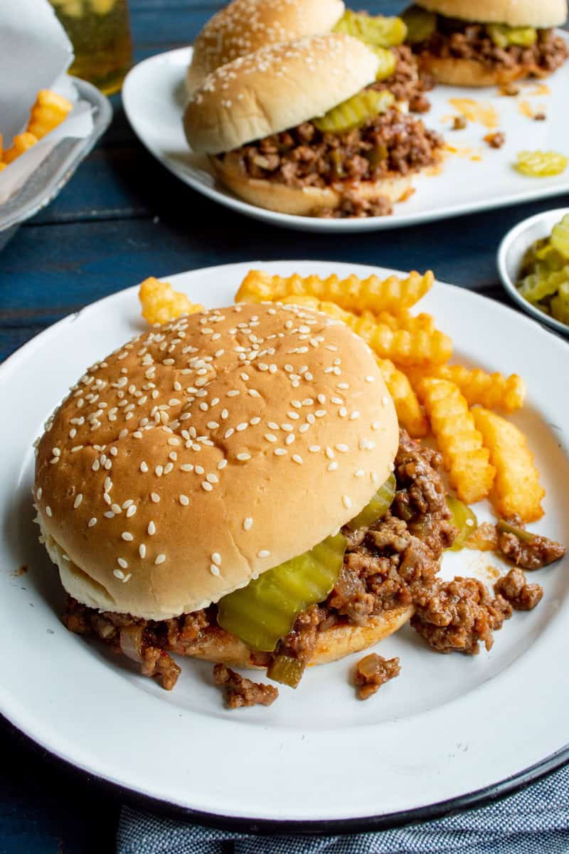 Heinz sloppy joe recipe with saucy ground beef and pickles on sesame seed buns, served with golden crinkle-cut fries on white plates.